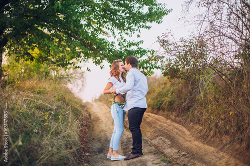 A loving couple are standing in the field road with trees at sunset, holding hands and kissing each other. Bright sunlight shines. Around the greens. Walk on the nature. Country style. © nataliakabliuk