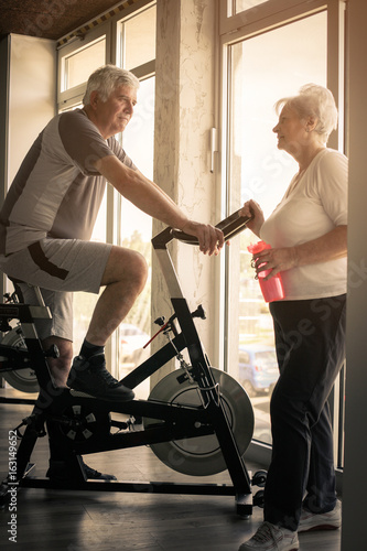 Two senior people at the gym. Senior man sitting on the elliptical machine and having conversation with senior woman.