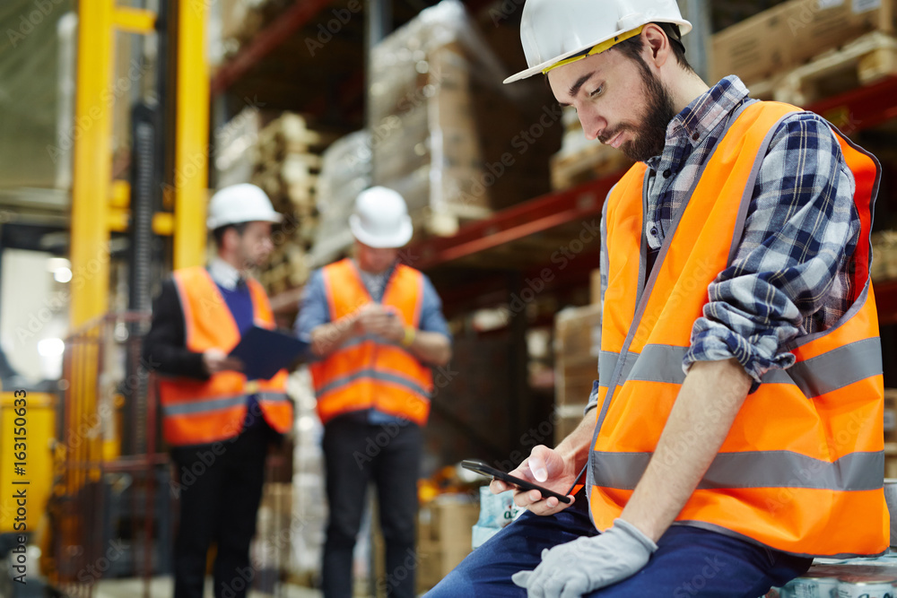 Man in uniform and helmet messaging at break in warehouse Man in uniform and helmet messaging at break in warehouse