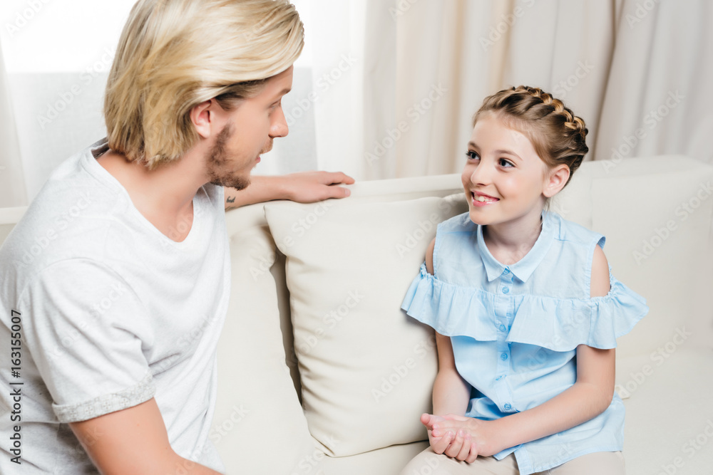 Fototapeta premium smiling father and daughter looking at each other and talking while sitting on sofa at home