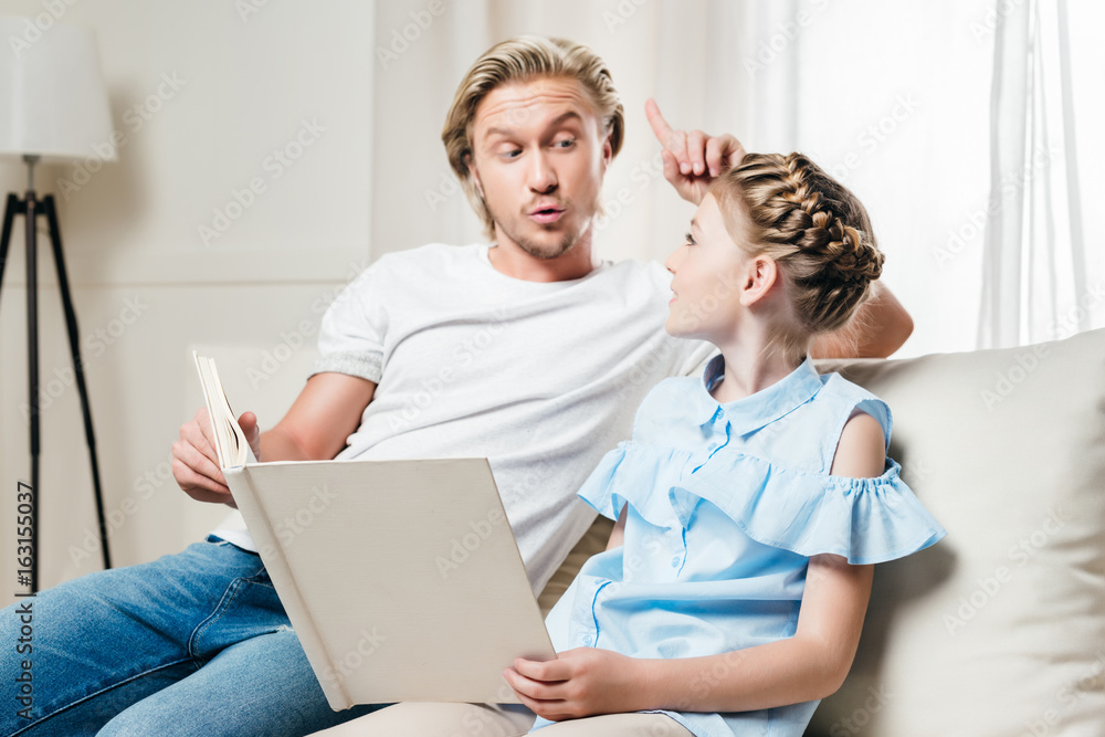 Obraz premium father pointing up while his daughter reading book and sitting on sofa at home