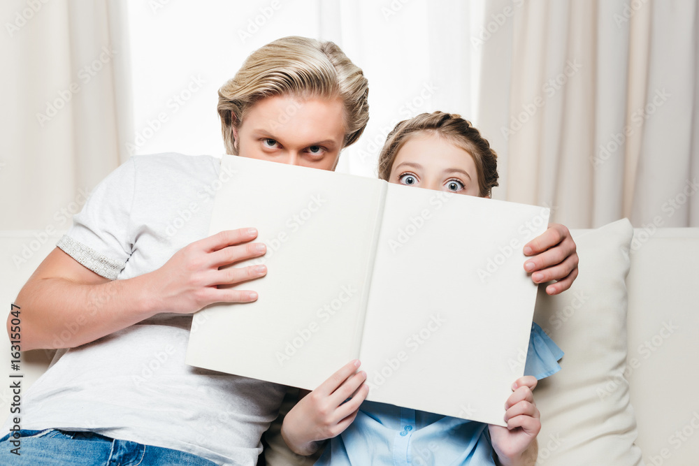 father and shocked daughter covering face with book while sitting on sofa at home father and shocked daughter covering face with book while sitting on sofa at home