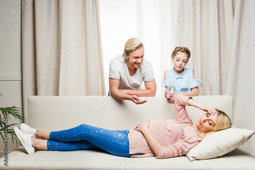 Smiling man and little girl holding medicine for pregnant woman lying on sofa