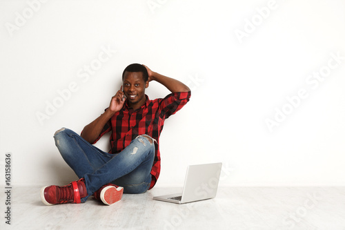 Happy black man using mobile and laptop sitting on the studio floor