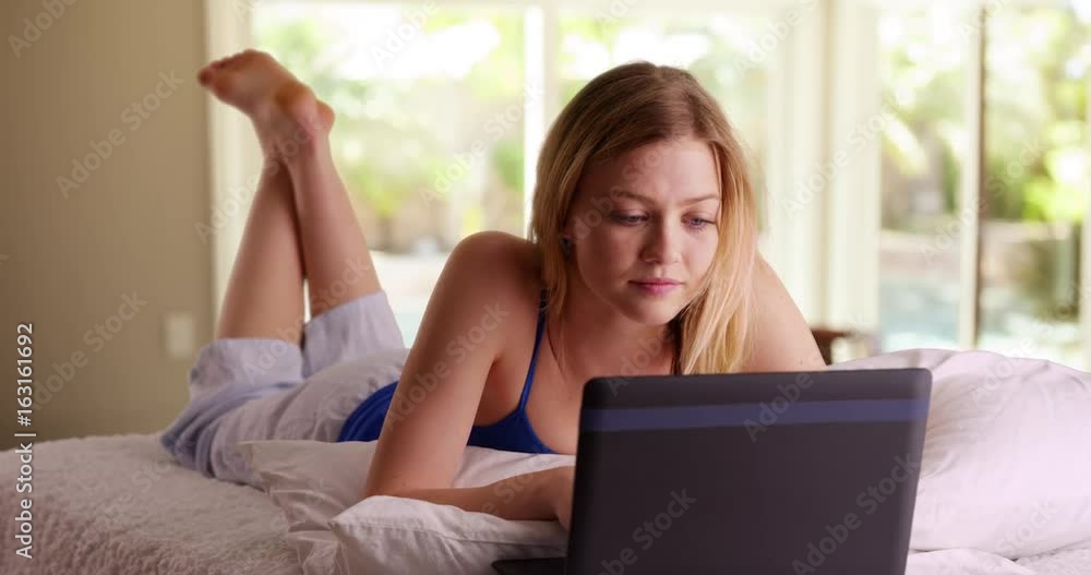 Young blonde millennial girl lying on bed using laptop computer Young blonde millennial girl lying on bed using laptop computer