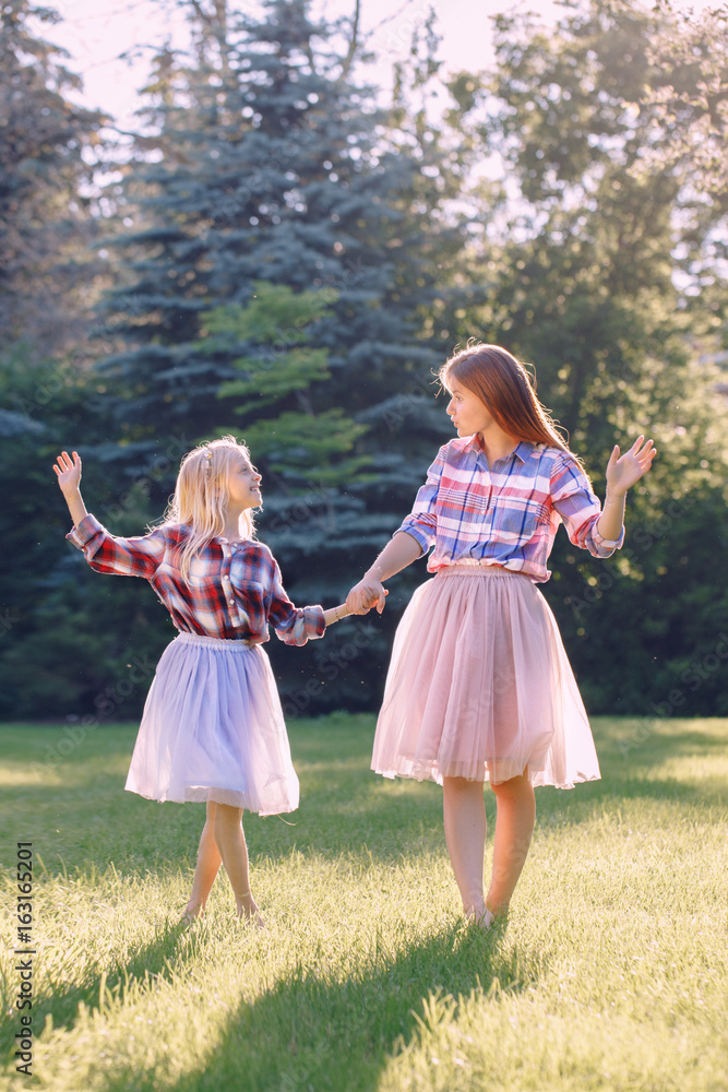 Naklejka premium Portrait of two smiling funny Caucasian girls sisters in plaid shirt and pink tutu tulle skirt, standing dancing barefoot on grass in park forest meadow at sunset. Friends having fun together.