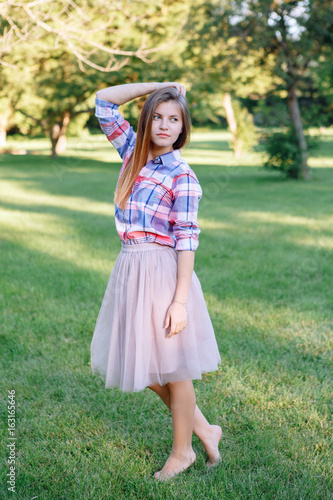 Portrait of beautiful young Caucasian woman with long red hair in plaid shirt and pink tutu tulle skirt, standing posing barefoot on toes on grass in park meadow at summer sunset, looking away