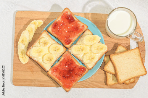 Square toast with jam and banana on a blue plate on a wooden board and a mug of milk