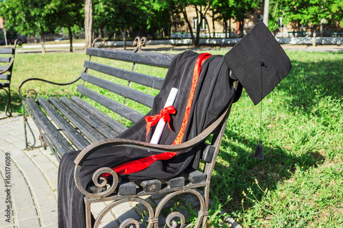 Graduation cap, hat with black tassel, mantle with a degree of paper on a park bench photo