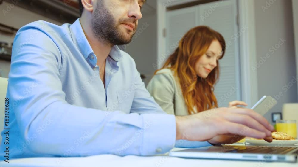 Side view of caucasian handsome young man with beard using laptop siting in kitchen with his wife using tablet. Indoor. Side view of caucasian handsome young man with beard using laptop siting in kitchen with his wife using tablet. Indoor.
