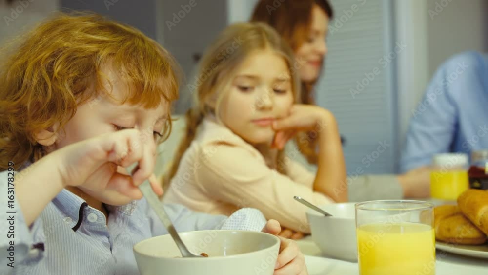 Caucasian young family of father, mother, dauther and little son having breakfast in kitchen. Little boy eating flakes with milk. Caucasian young family of father, mother, dauther and little son having breakfast in kitchen. Little boy eating flakes with milk.