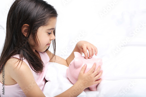 Beautiful little girl with piggy bank on white bed