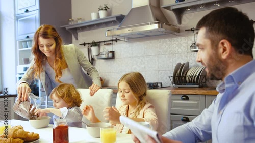 Caucasian little boy, girl and father siting on the table in kitchen. Father reading newspaper, mother filling flakes in plate of son, dauther eating flakes with milk. photo