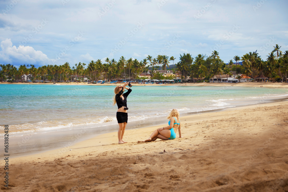 girl traveller making the photo of another girl in bikini on the sandy beach