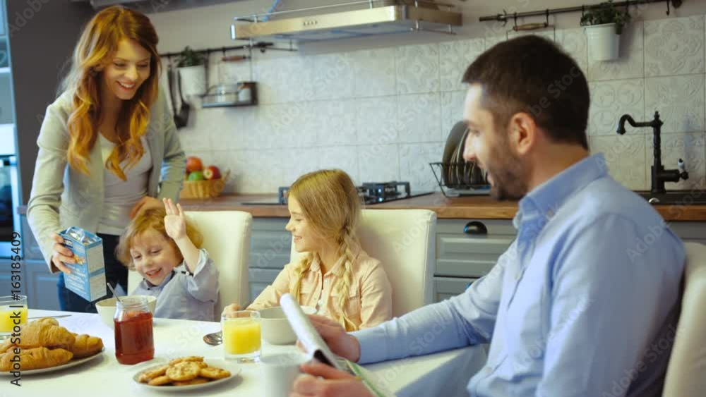 Caucasian young family in kitchen. Caucasian attractive woman filling flakes in children plates while father reading newspaper. Caucasian young family in kitchen. Caucasian attractive woman filling flakes in children plates while father reading newspaper.