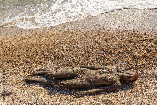 Beautiful woman laying on the beach and sea waves splash near her.Top view. Woman smearing mud mask on body, Spa outdoor and cosmetics. Recreation tourism and lifestyle, body care, natural therapy.