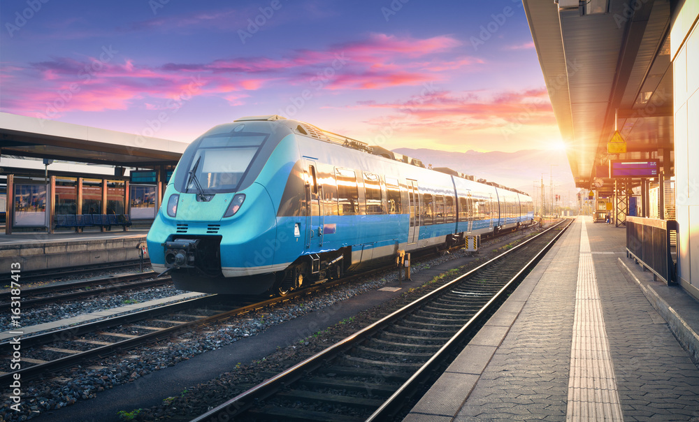 Naklejka premium Nowoczesny pociąg podmiejski wysokiej prędkości na stacji kolejowej i kolorowe niebo z chmurami o zachodzie słońca w Europie. Przemysłowy krajobraz z błękitnym pociągiem pasażerskim na kolejowej platformie. Tło kolejowe