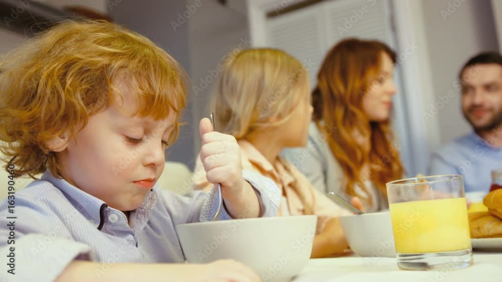 Portrait of caucasian beautiful little boy with blue eyes and blonde hair eating flakes with milk in kitchen with his family. Portrait of caucasian beautiful little boy with blue eyes and blonde hair eating flakes with milk in kitchen with his family.