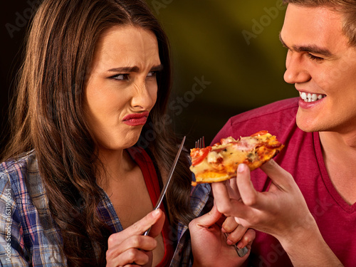 Couple eating in fast food restaurant. Man and woman eat slice pizza with knife and fork. Friends holding junk on dark background. Girl against the use of semi-finished products idea.