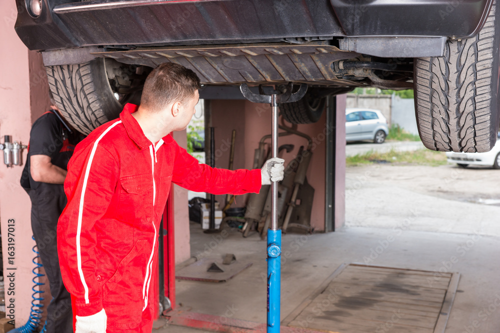 Fototapeta premium Young auto mechanic in uniform holding equipment for elevating car on a lift
