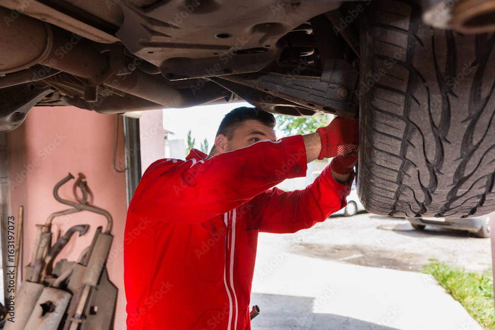 Car mechanic in uniform is repairing wheel while working underneath a lifted car