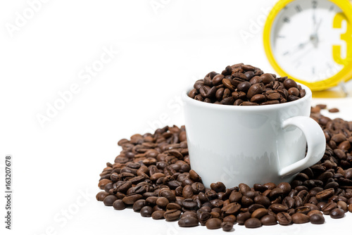 cup with coffee beans and yellow alarm clock on white background  closeup