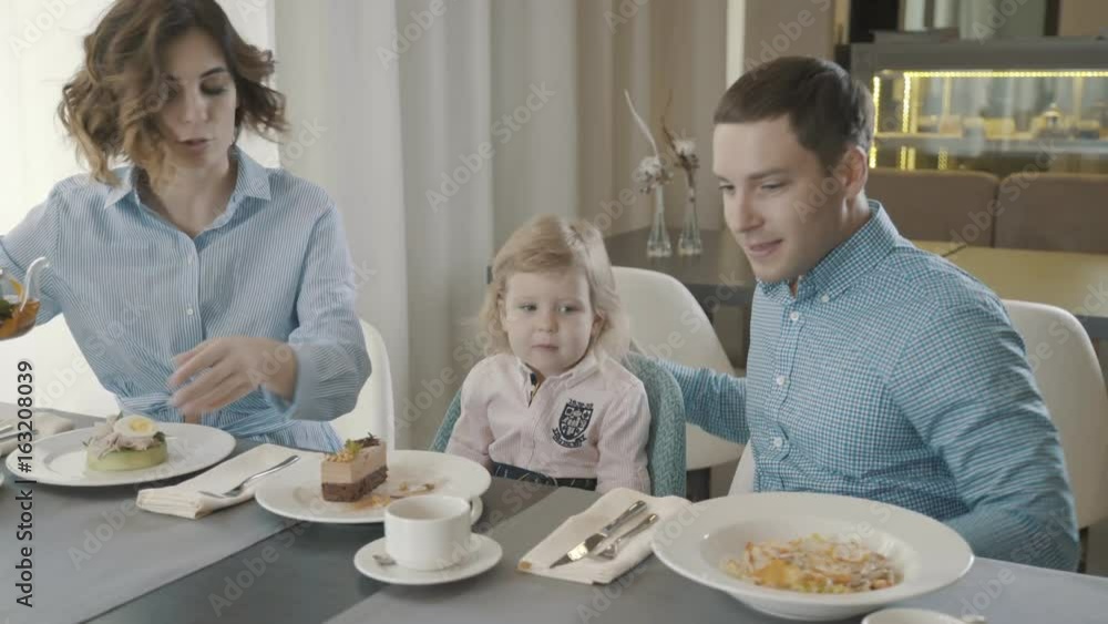 A family with a small child in a restaurant: dad, mom and daughter eat breakfast in a cozy family cafe, a woman pours tea in white mugs. Family Idyll, Morning Tea Party. The whole family is in shirts. A family with a small child in a restaurant: dad, mom and daughter eat breakfast in a cozy family cafe, a woman pours tea in white mugs. Family Idyll, Morning Tea Party. The whole family is in shirts.