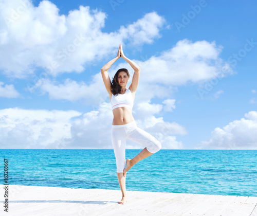 Young and beautiful girl in white sportswear doing yoga on a wooden pier at summer. Yoga, sport, leisure, vacation and traveling concept.
