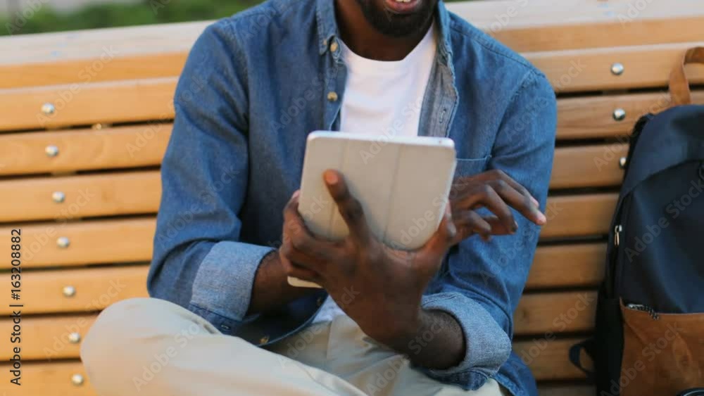 Beautiful young african man in glasses smiling, using tablet for chatting with friend, siting on the bench in park. Beautiful young african man in glasses smiling, using tablet for chatting with friend, siting on the bench in park.
