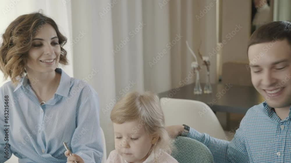 Modern family with a small child in a family restaurant: a blond pretty girl is happy to be with her parents. Parents are touched by their daughter, who tries to eat by herself. Modern family with a small child in a family restaurant: a blond pretty girl is happy to be with her parents. Parents are touched by their daughter, who tries to eat by herself.