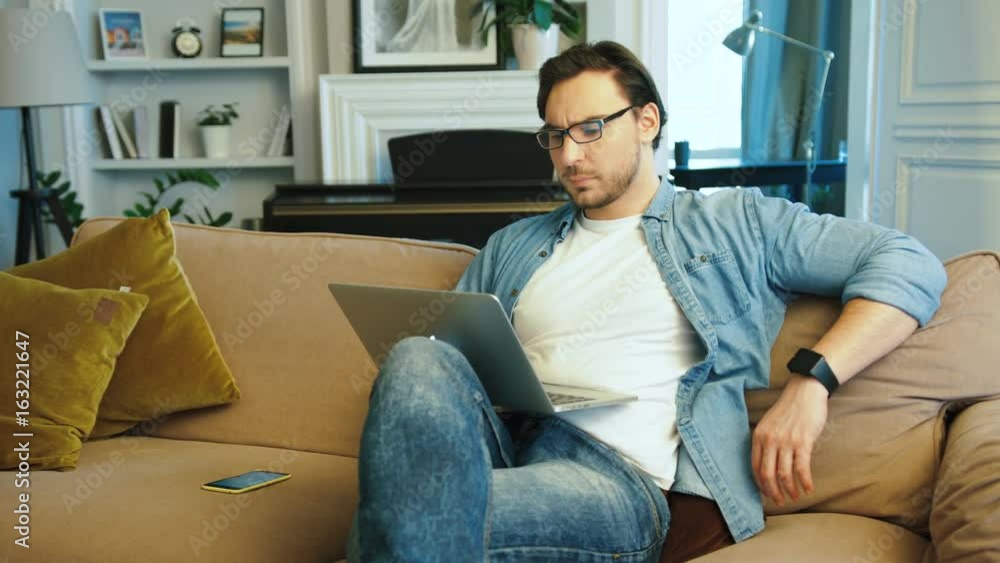 Young tired man in casual blue shirt with glasses working on the laptop while sitting on the sofa at home. Young tired man in casual blue shirt with glasses working on the laptop while sitting on the sofa at home.