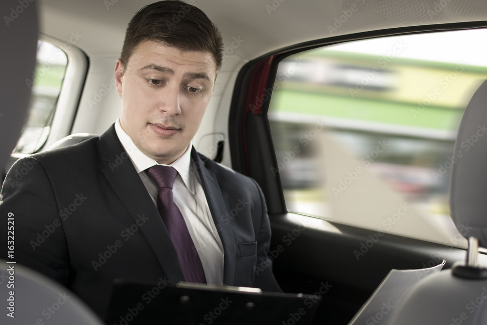 Surprised businessman working while sitting in a car. Man beeing driven to work in his limo. Suit and tie businessman in the back seat while the limo driver is driving.