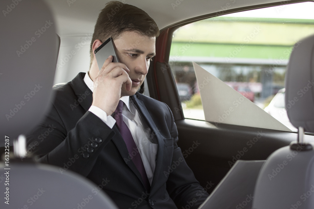 Businessman working while sitting in a car. Man beeing driven to work in his limo. Suit and tie businessman in the back seat making a call while the limo driver is driving. Businessman working while sitting in a car. Man beeing driven to work in his limo. Suit and tie businessman in the back seat making a call while the limo driver is driving.
