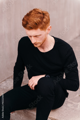 Thoughtful young man sitting on pavement in the street. Red hair handsome male model. Outdoor. Wearing black