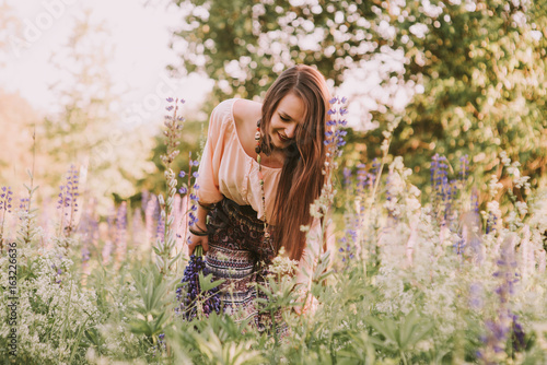 Young beautiful brown hair hippie girl gathering flowers and walking through meadow in sunny day. Summer field. Great force of unity with the Nature. Outdoors