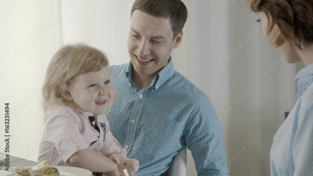 A little girl is sitting on her father's lap, Mom is playing with her, holding a soft toy in her hand. A happy child plays with his parents: the girl joyfully screams and rejoices. A family of three A little girl is sitting on her father's lap, Mom is playing with her, holding a soft toy in her hand. A happy child plays with his parents: the girl joyfully screams and rejoices. A family of three