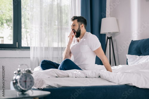 Tableau sur toile Bearded young man in pajamas yawning while sitting on bed in morning