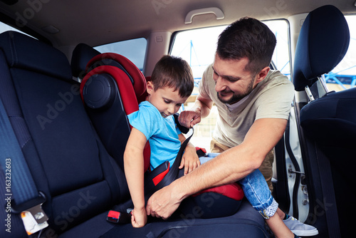 Concentrated father helps his son to fasten belt on car seat