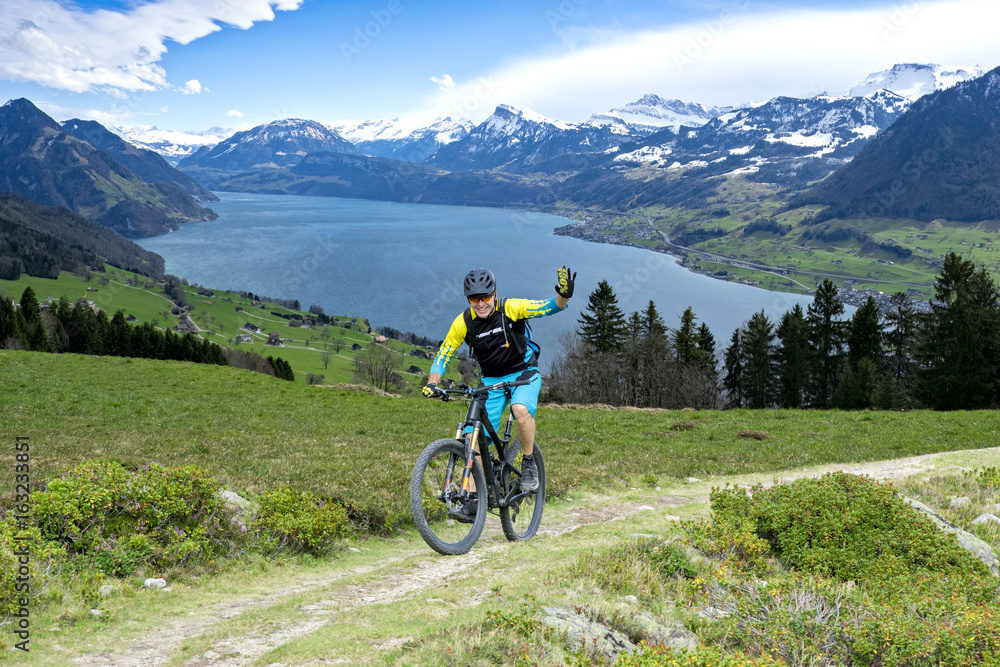 Sportive man in middle age with mountain bike on mountain trail beckons the viewer. The background shows the Lake Lucerne.