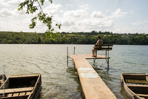 Fototapeta Naklejka Na Ścianę i Meble -  Man sitting on pier, wooden boat on river background