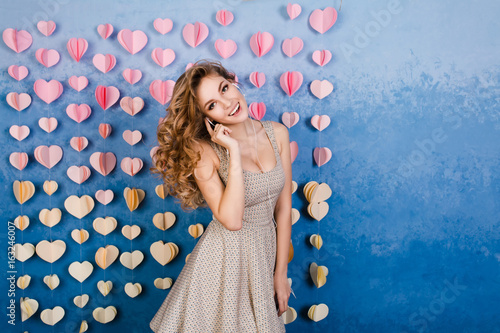 Cute sexy slim girl with blond curly hair standing in a studio with blue background and listenign to music on earphones. She smiles and looks sexy. photo