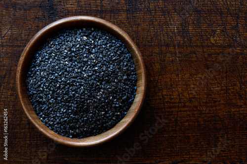 Black sesame seeds in dark wooden bowl isolated on dark brown wood from above.