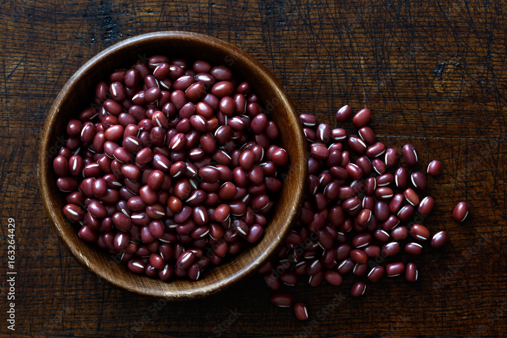 Dry adzuki beans in dark wooden bowl isolated on dark brown wood from above. Spilled beans. Dry adzuki beans in dark wooden bowl isolated on dark brown wood from above. Spilled beans.