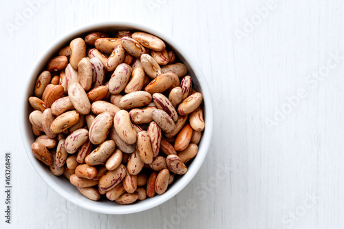 Dry pinto beans in white ceramic bowl isolated on painted white wood from above.