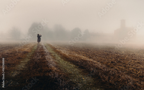 Unidentifable woman in the distance looking at a church in the dense fog of the Lombardy region of Italy during the winter, frozen corn field photo