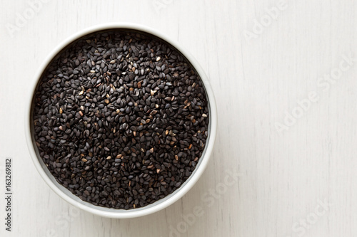 Black sesame seeds in white ceramic bowl isolated on painted white wood from above. 