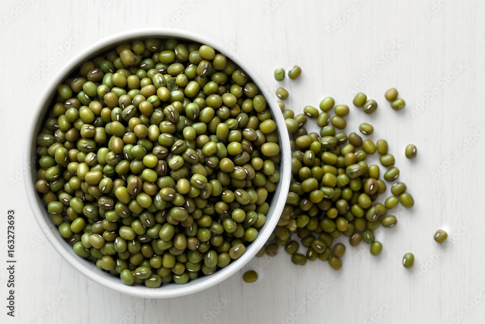 Dry mung beans in white ceranic bowl isolated on painted white wood from above. Spilled beans. Dry mung beans in white ceranic bowl isolated on painted white wood from above. Spilled beans.