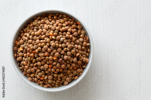 Dry unpeeled red lentils in white ceramic bowl isolated on painted white wood from above. photo