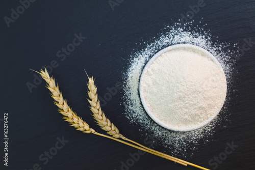 Wheat flour in white cup decorate by wheat ear on black slate stone plate with copy space for preparing baking or pasta