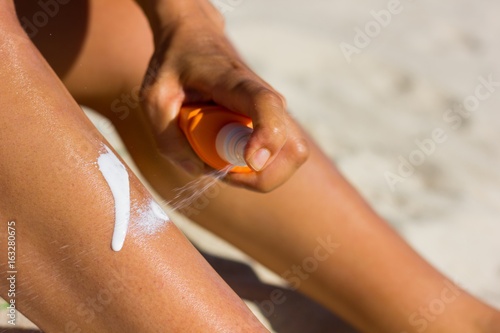 Woman applying sun lotion on her leg with a spray at the beach on a hot, sunny day